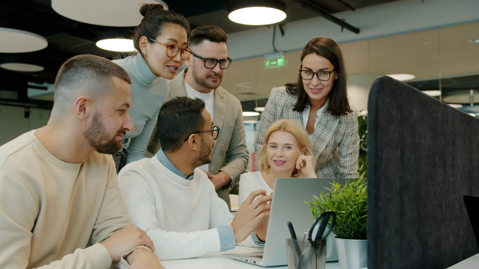 A team of coworkers gathers around a laptop in a bright open-plan office.