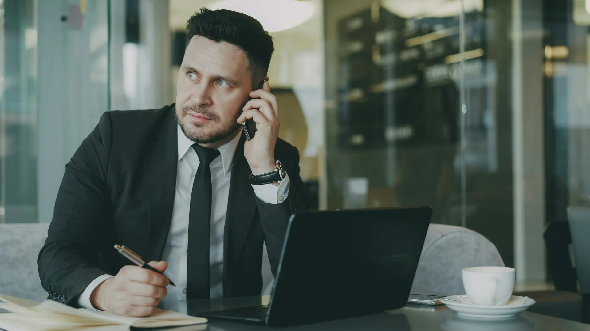 A businessman in a suit takes a phone call at his laptop.