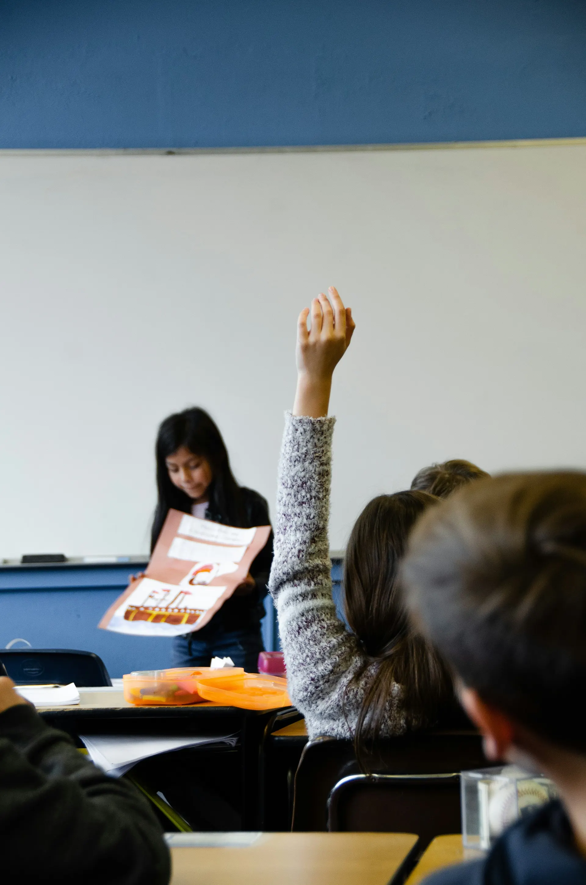 Students in a classroom, one raising a hand while another holds a handout.