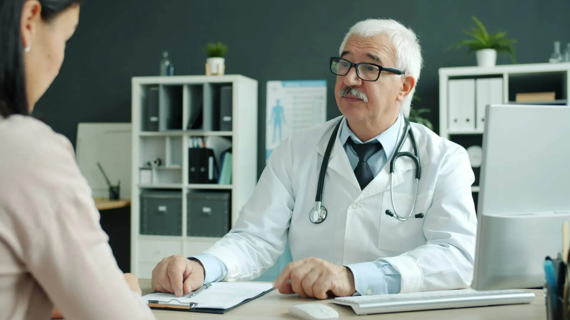A doctor in a white coat talks with a patient across his desk.