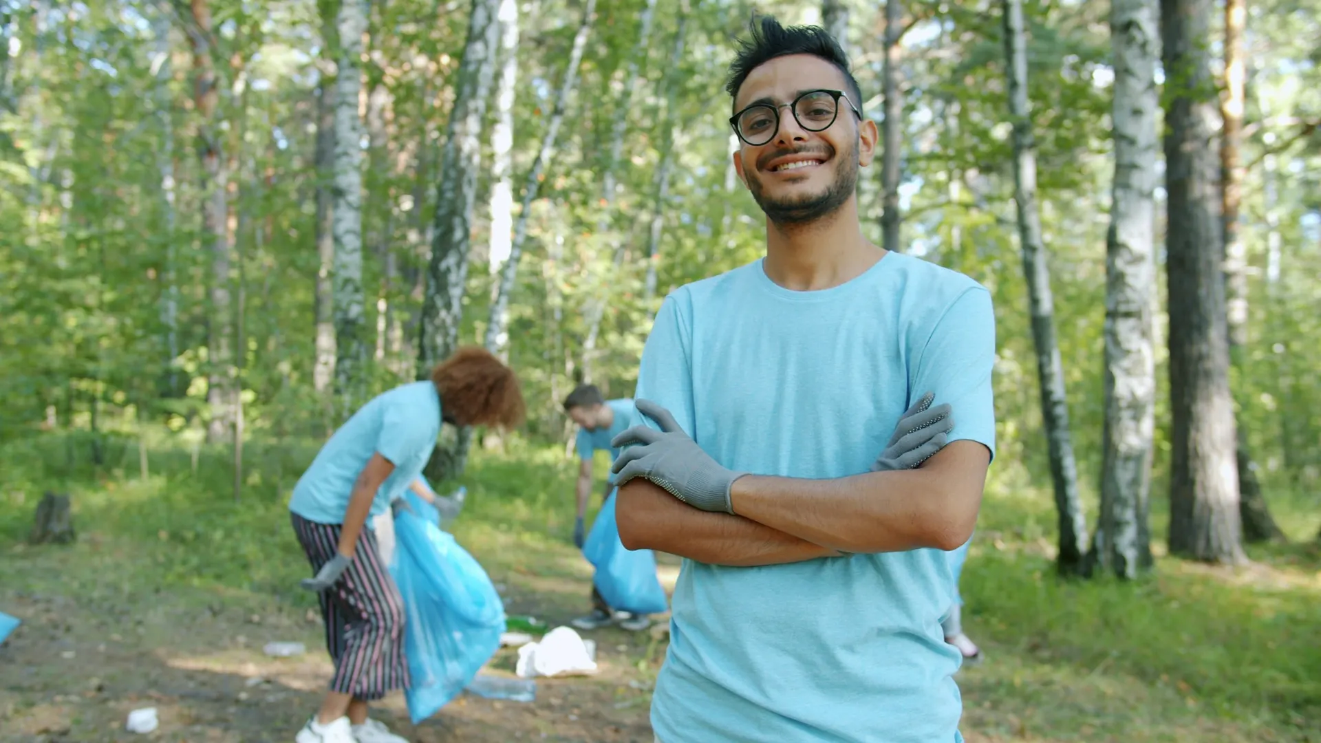 A smiling volunteer stands in a park while others collect litter behind him.